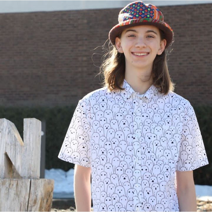 Image of Wesley standing outside in front of Northland Community and Technical College. He is wearing a white button down shirt with ghosts on it. He has shoulder length brown hair. He is wearing a hat that is make up of colorful patches of fabric. The hat is flat on the top and has a short round brim.